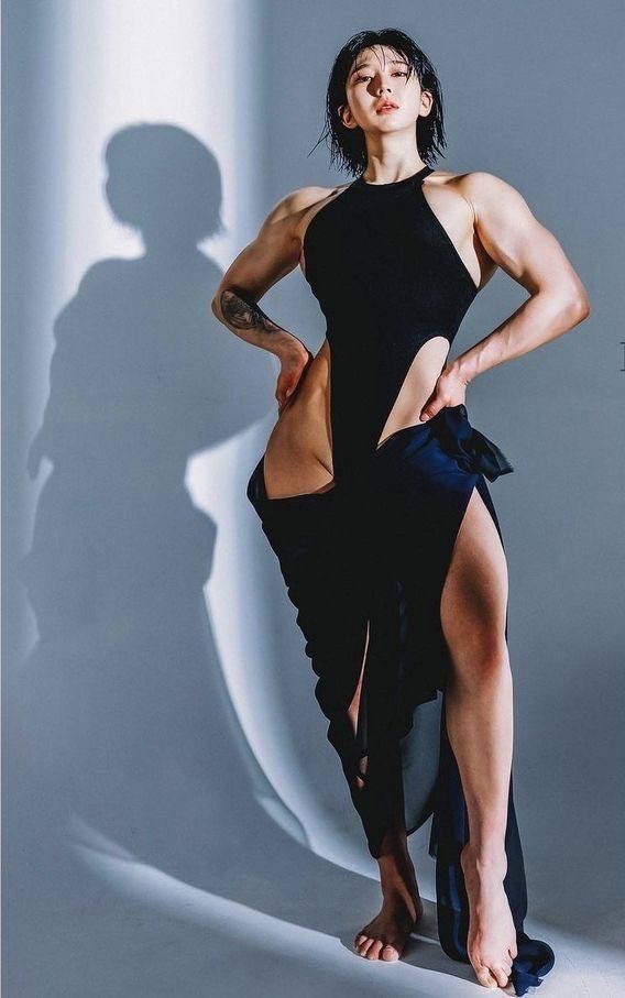 a muscular woman in a black bodysuit poses against a light grey background. she is standing with one leg slightly forward and her hands on her hips, showcasing her toned physique. the studio lighting casts dramatic shadows on the wall behind her.