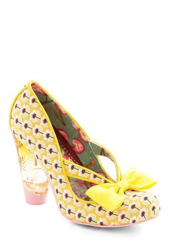 a closeup shot of a single irregular choice women's shoe in yellow. the shoe features a high platform heel, a floral pattern, and a bright yellow bow. the heel is transparent and pastel pink at the bottom. the shoe is set against a white background.