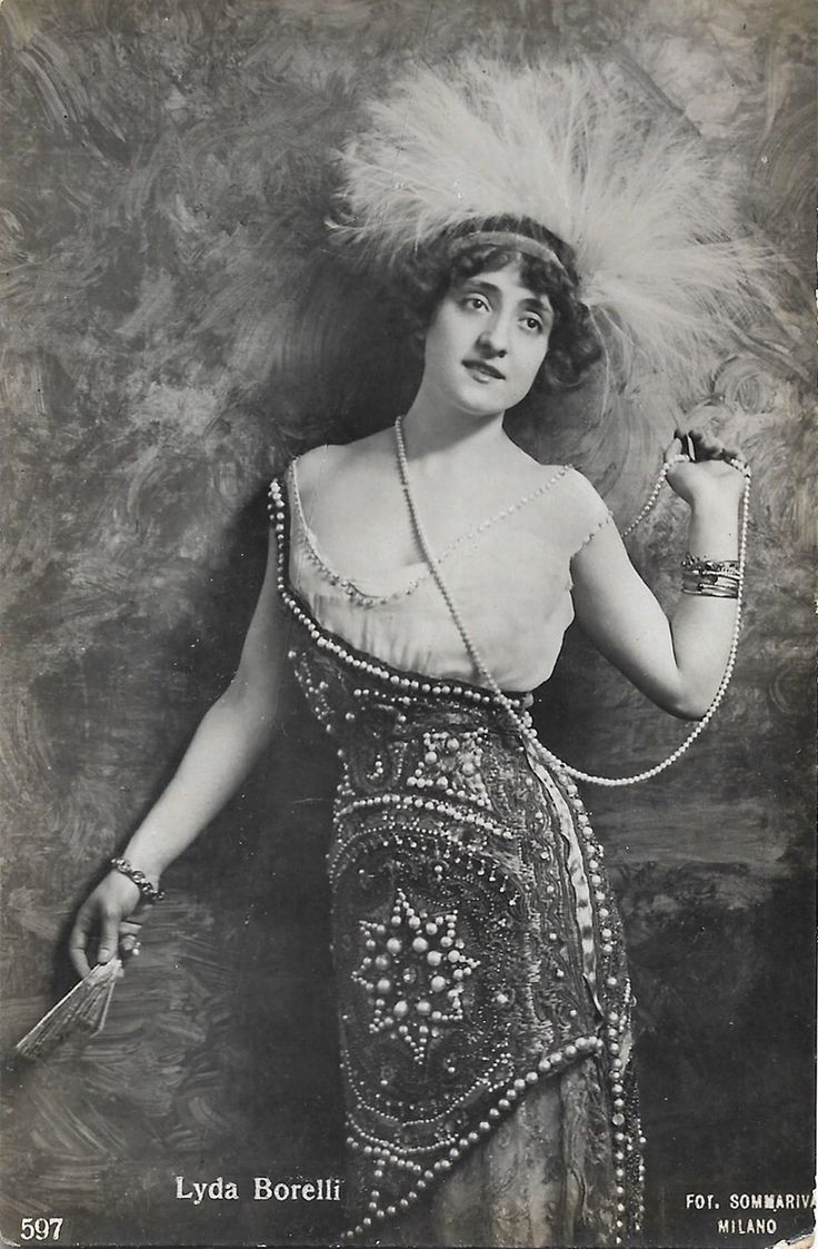 a vintage black and white portrait of lyda borelli, an actress. she is wearing a dress heavily decorated with pearls, a large feathered headdress with a headband, and holding a string of pearls in her hand. she also has bracelets on both wrists and is holding a fan in her left hand. the photograph is attributed to fot. sommariva milano.