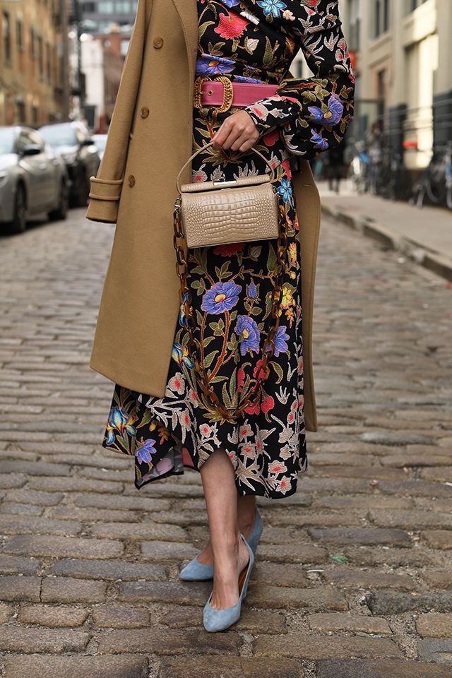 a stylish woman walks down a cobblestone street in the city. she's wearing a floral midi dress, a camelcolored coat, blue suede pointedtoe heels, and a beige crocodileembossed crossbody bag. her outfit is accessorized with a pink belt and subtle jewelry. the overall aesthetic is elegant and sophisticated. the image showcases a fall or autumnal urban street style.