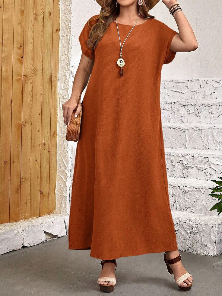 a woman is standing in front of a wooden wall wearing a rustcolored maxi dress. the dress is loosefitting, with short sleeves and a round neckline.  she is accessorized with a straw bag, sandals, and a necklace.  the overall style is casual and bohemian. the image is welllit and shows the woman from head to toe.