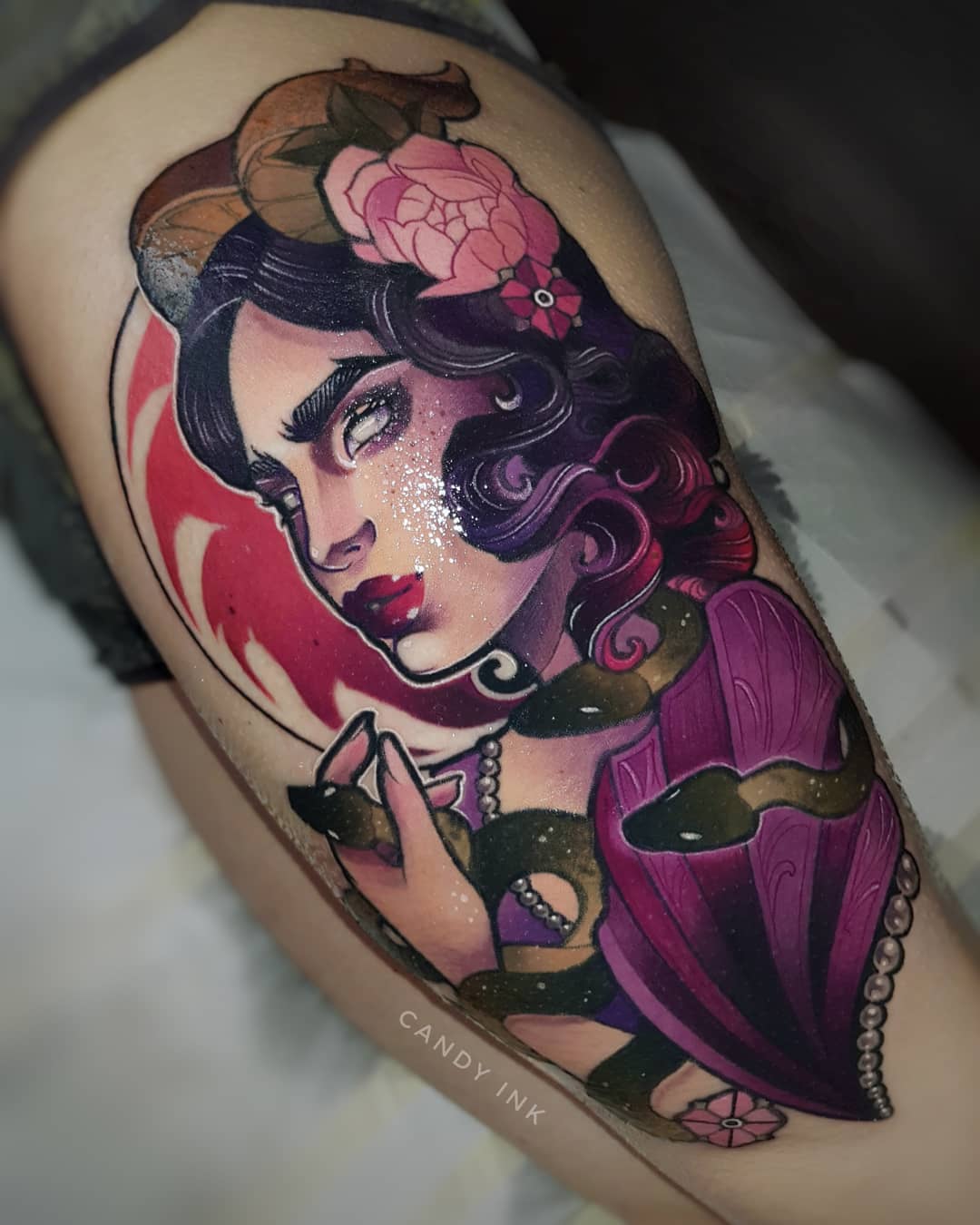 a neotraditional tattoo on someone's upper arm. it depicts a woman from the chest up with purple hair, horns, and snakes. her skin is fair and there is a red crescent moon shape behind her head. she is wearing a deep purple garment with pearl accents. she has flowers in her hair and near her arm. the style of the tattoo is neo traditional and very colorful.