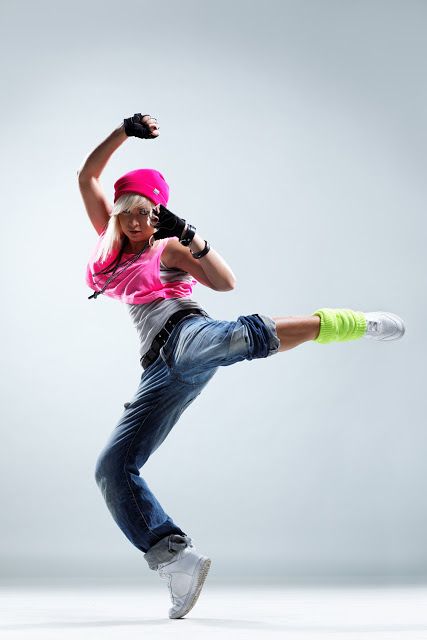 a young woman in a pink hat and jeans is middance pose. she is facing to the left and her right leg is lifted, bent at the knee, and extended to the right. her left leg is bent at the knee, with the toes touching the ground. her arms are extended, one above and one below her head.  she is in a studio setting.