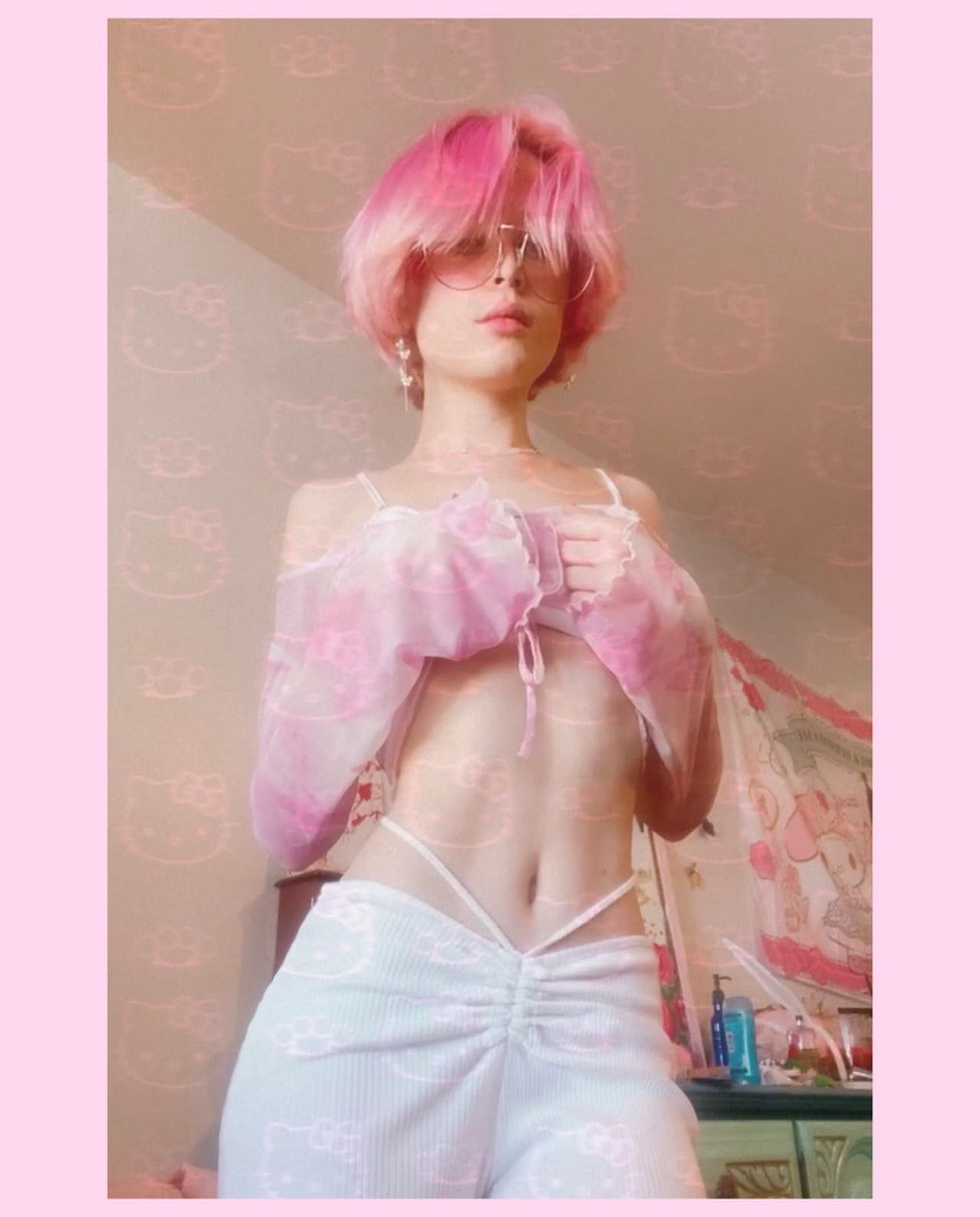 a young woman with short, bright pink hair is posing in a pastel pink and white outfit. she's wearing a sheer, tiedye cropped top and lowrise white pants with a drawstring waist. the pants and top feature a subtle hello kitty print.  she's also wearing sunglasses and has a generally soft and feminine aesthetic. the background is a bedroom setting with pink wallpaper and a vanity table visible. the overall vibe is kawaii and cute.