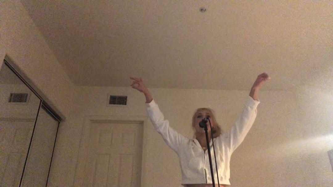 a woman with blonde hair stands in a room singing into a microphone with her arms raised. she is wearing a white jacket. behind her, there is a door, a closet, a light fixture on the ceiling, and a ventilation grille.