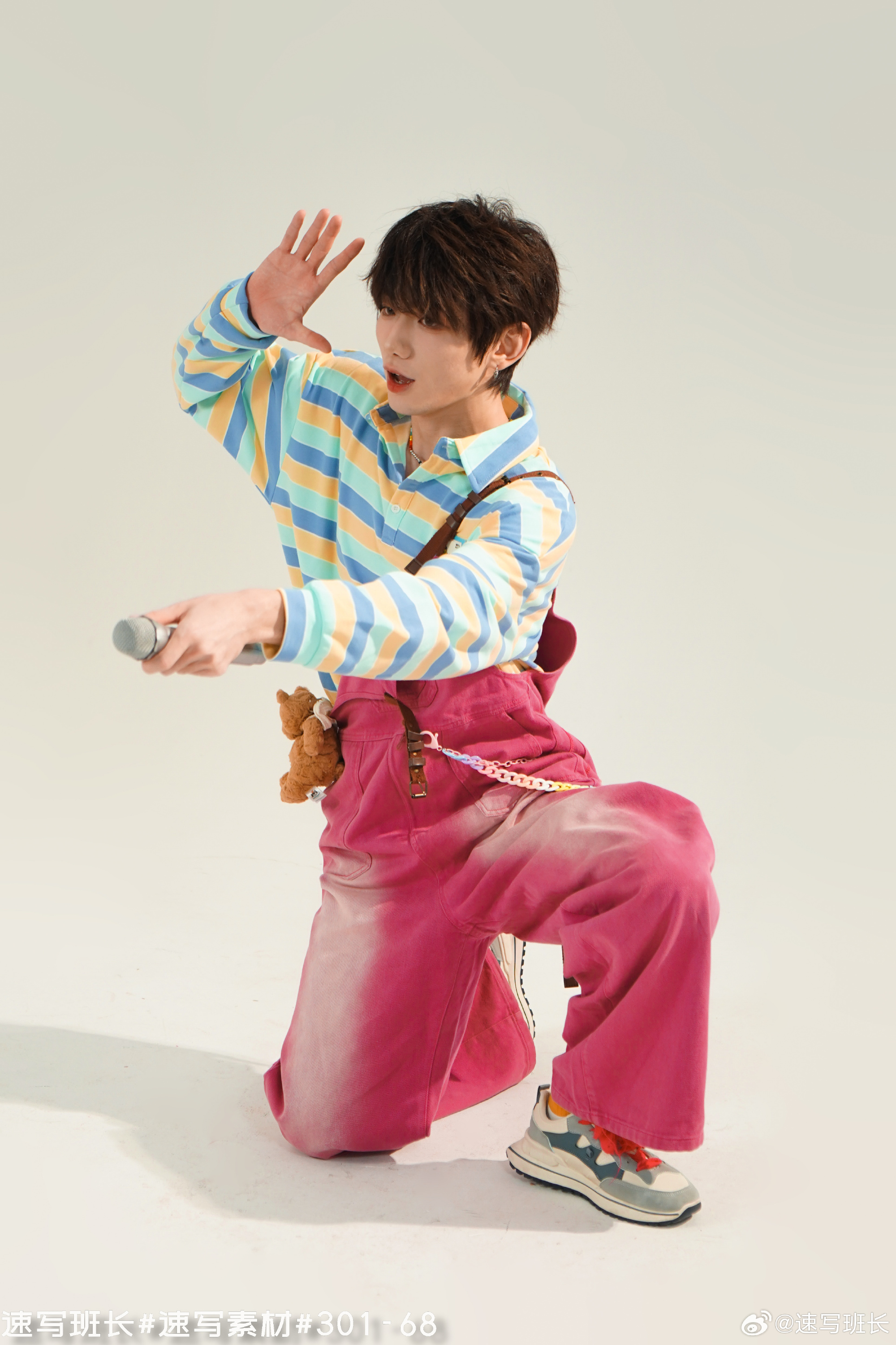 a young man wearing a colorful striped shirt, pink overalls, and sneakers is posing with a microphone in his hand, as if singing or rapping. he is crouched down, with one hand raised, against a minimalist white backdrop. the lighting is bright and artificial, likely a studio setting.