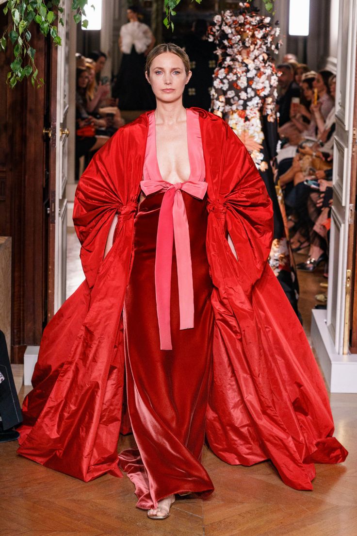 a model walks the runway wearing a stunning red velvet gown with a flowing cape. the gown features a plunging neckline and a large bow at the waist. the model's hair is pulled back, and she is wearing minimal makeup. the overall look is elegant and feminine, showcasing the beauty and craftsmanship of high fashion.