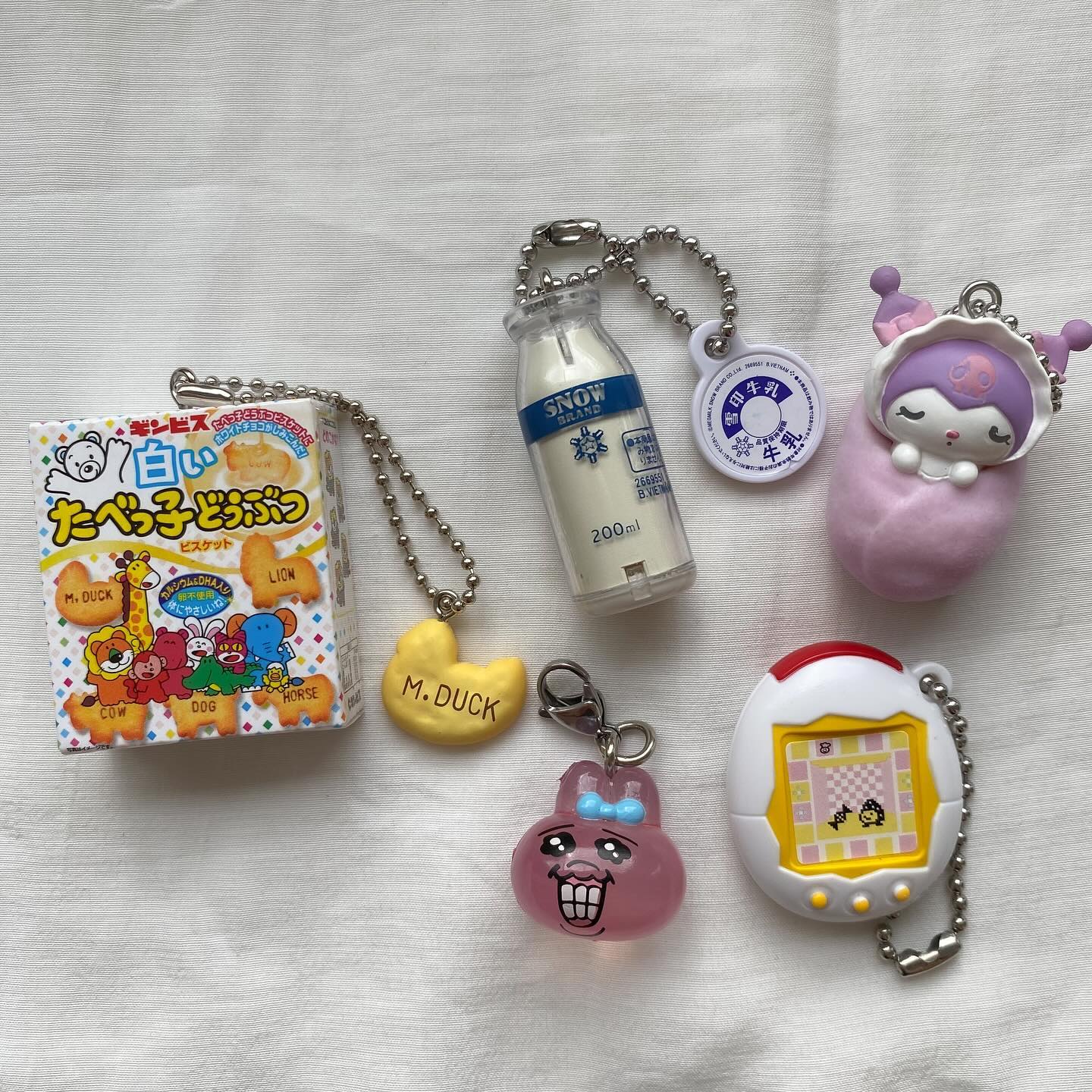 the image contains various keychains and charms. a package of ginbis animal cracker keychains is next to a snow brand milk bottle keychain. a yellow duckshaped keychain, a pink bunny keychain, a tamagotchi keychain, and a my melody charm.