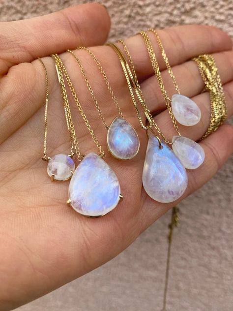 a closeup image showcasing a hand holding multiple delicate gold necklaces adorned with iridescent moonstone pendants. the moonstones vary in size and shape, ranging from small round cabochons to larger teardrop cuts. the soft, natural lighting highlights the ethereal glow of the stones against the warm tone of the skin.