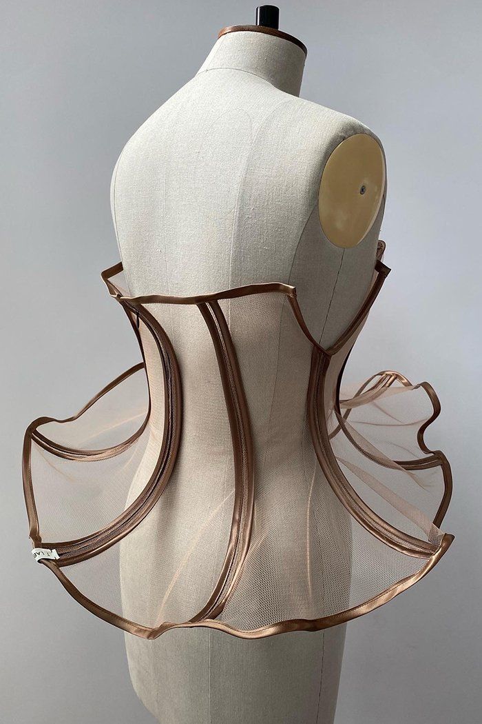 a photograph shows a mannequin wearing a corsetlike garment. the mannequin is beige with a brown trim at the top and a black knob on top of the head. the corset has a frame with brown trim and beige mesh between the trim. the trim creates curved shapes that flair out at the hips.