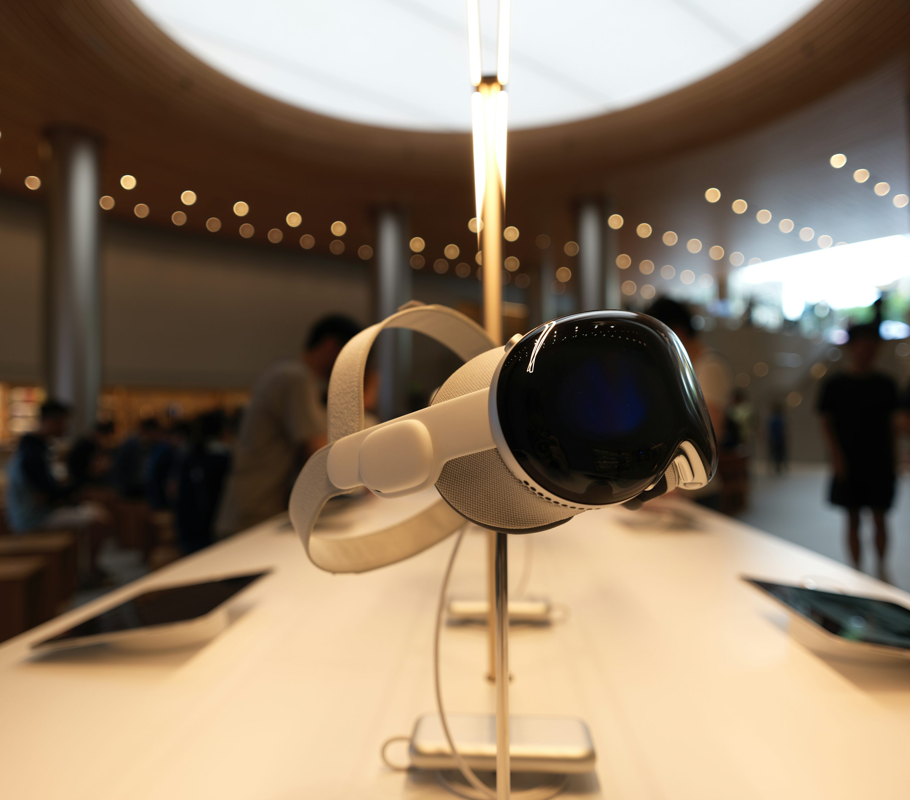 a white vr headset is displayed on a white table in a store. the headset is on a stand, and there are other devices on the table as well. people are in the background. the store appears to be an apple store, based on the interior design and layout.