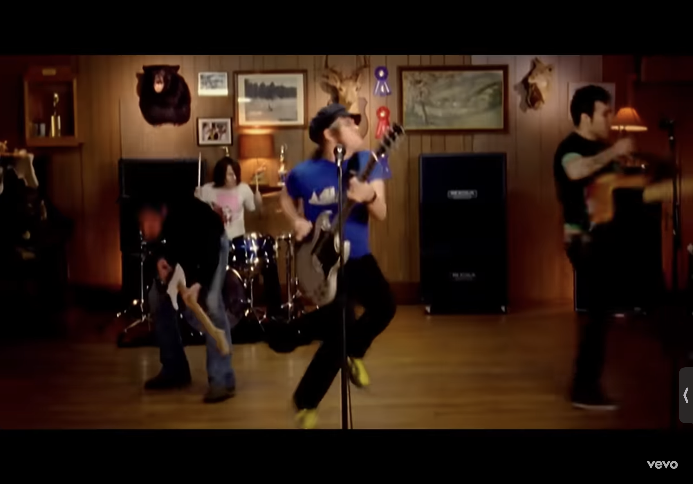 the image shows a rock band performing in a woodpaneled room. the lead singer, wearing a blue tshirt and a black beanie, is front and center, energetically playing a white electric guitar. he is flanked by two other guitarists, a drummer in the background, and what appears to be a female backing vocalist. the room resembles a cabin with mounted animal heads and a rustic ambiance. the lighting is bright and stagelike, focused on the band members.  the overall impression is one of highenergy, live rock music.
