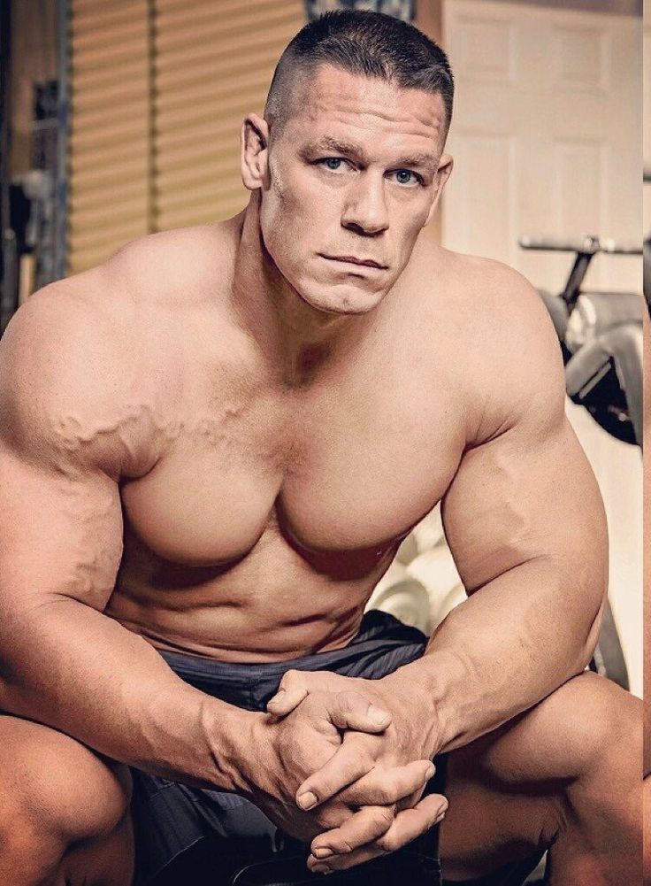 a closeup shot of john cena, a famous wwe wrestler, sitting in a gym setting. he's shirtless, showcasing his incredibly muscular physique with prominent veins visible throughout his arms and chest. his expression is serious and focused, hands clasped together in his lap. the background is slightly blurred but shows gym equipment, suggesting a workout or training environment.  the image highlights his powerful build and intense demeanor.