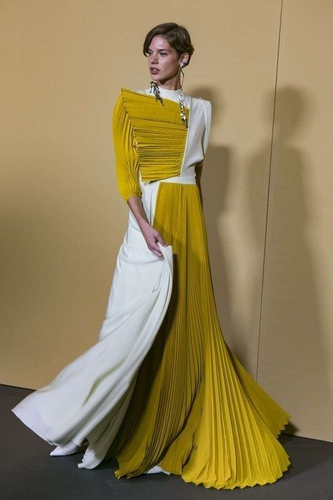 a woman model wears a long, pleated yellow and white dress, the top half is white and the bottom is yellow, both with pleats. she poses in a studio with the background a neutral yellow, she is wearing a pair of earrings and has her hair pulled back from her face. the dress is very elegant and stylish.