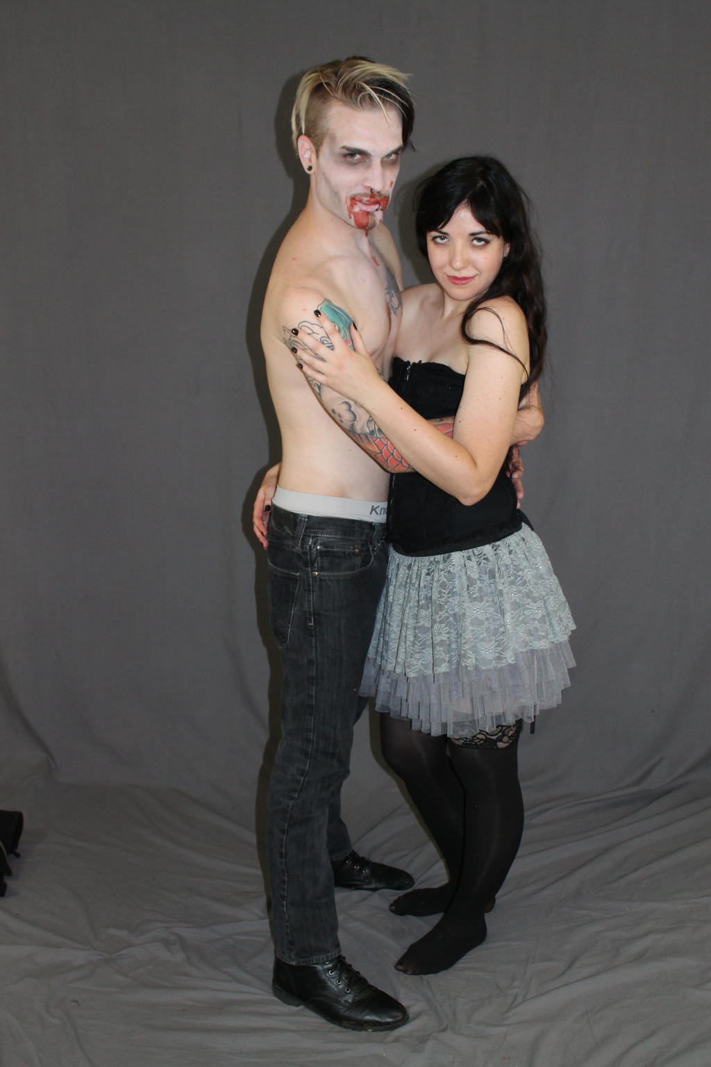 a full shot of a couple, a man and a woman, posed together against a grey backdrop. the man is shirtless, showing off tattoos on his arms, and his face has vampirelike makeup applied. his hair is blonde. the woman is wearing a grey tutulike skirt and black stockings. she is wearing black eyeliner and has dark hair. they appear to be dressed in goth or vampirethemed costumes.  they are embracing, and the woman has her arm around the man's waist.  they are both looking at the camera.