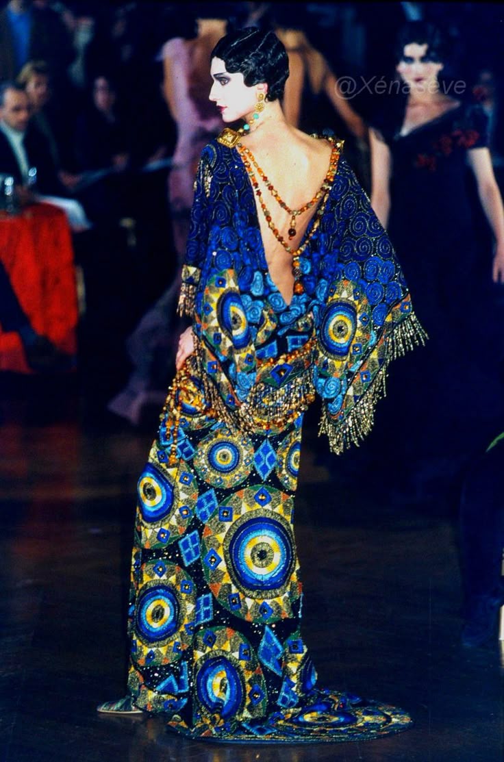 a model is walking on a runway wearing a long, blue, openback dress with beaded embellishments. the dress features a repeating pattern of circles and diamond shapes, and is adorned with strands of beads. the model is wearing matching beaded earrings and a hairstyle reminiscent of the 1920s. there are spectators in the background.