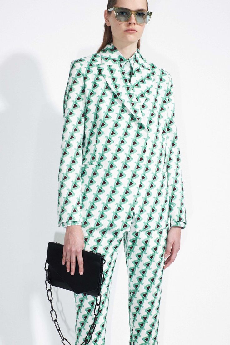 a fashion model is showcasing a stylish green and white geometric print suit. the suit consists of a blazer and matching pants. she is also carrying a black chain bag. the model is wearing sunglasses and has her hair pulled back. the backdrop is a plain white wall. the overall style is contemporary and chic.
