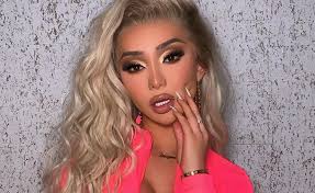 closeup portrait of a young woman with long, wavy blonde hair. she is wearing a vibrant neon pink jacket and has noticeable makeup, including dark eyeshadow and pink lipstick. she is accessorized with earrings, a bracelet, and rings, and her nails are manicured. her expression is confident and stylish, possibly a professional headshot or promotional image.  the background is a mottled gray textured wall.
