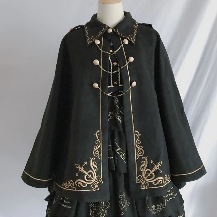 a black lolita fashion cape with gold embroidery and buttons, worn over a dress. the cape has a collar and a chain detail. the dress is also black with white designs.