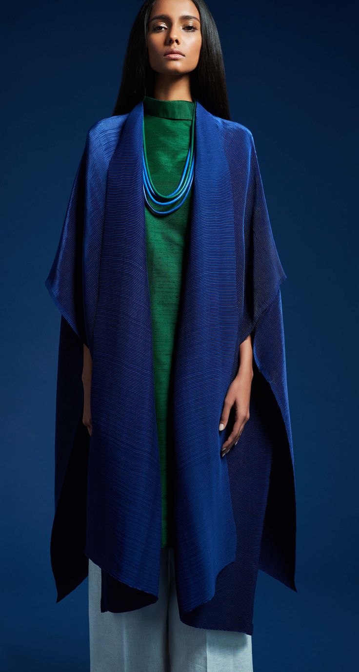 a woman model is standing against a dark blue background. she is wearing a long blue pleated coat over a green dress. the coat is open in the front, and the dress is visible underneath.  she is wearing a multilayered blue necklace. the woman has dark hair and is looking directly at the camera.  the image is a fashion shot with a focus on the clothing.