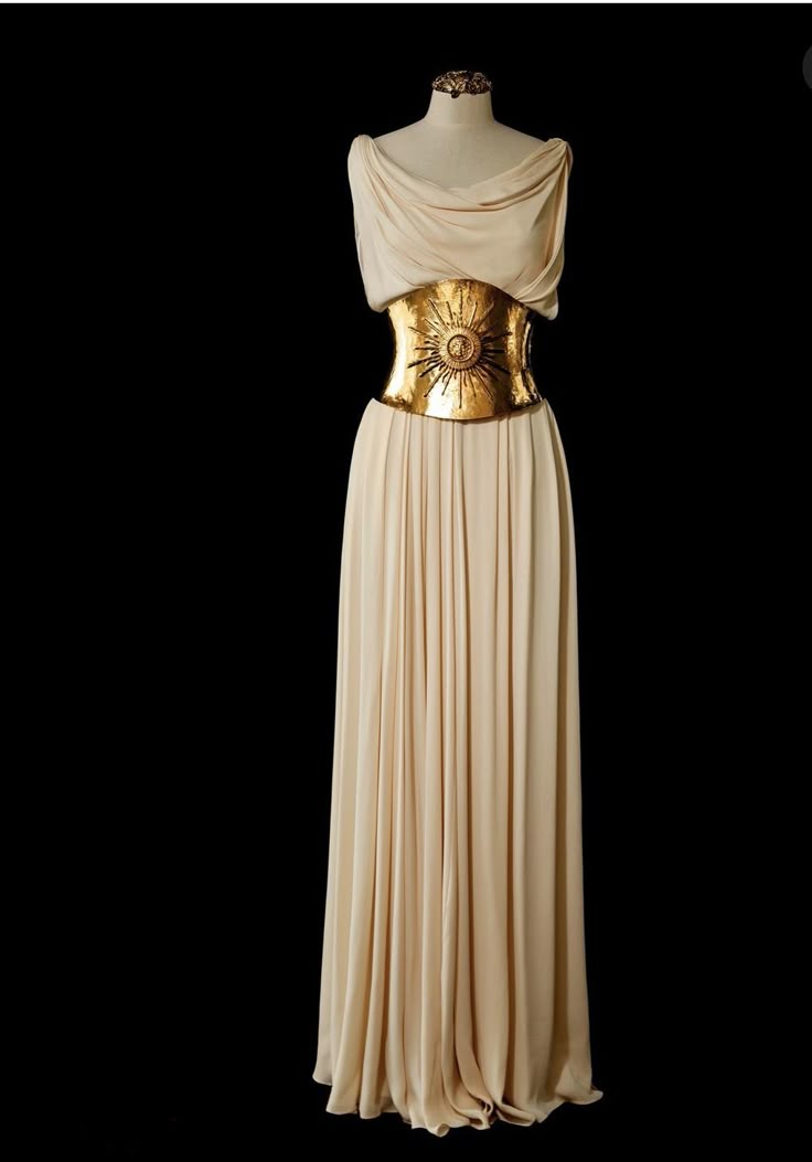 a creamcolored floorlength gown is displayed on a mannequin against a black backdrop. the gown features a draped bodice and a pleated skirt. a wide, ornate gold belt with a sun design cinches the waist, adding a striking visual element.