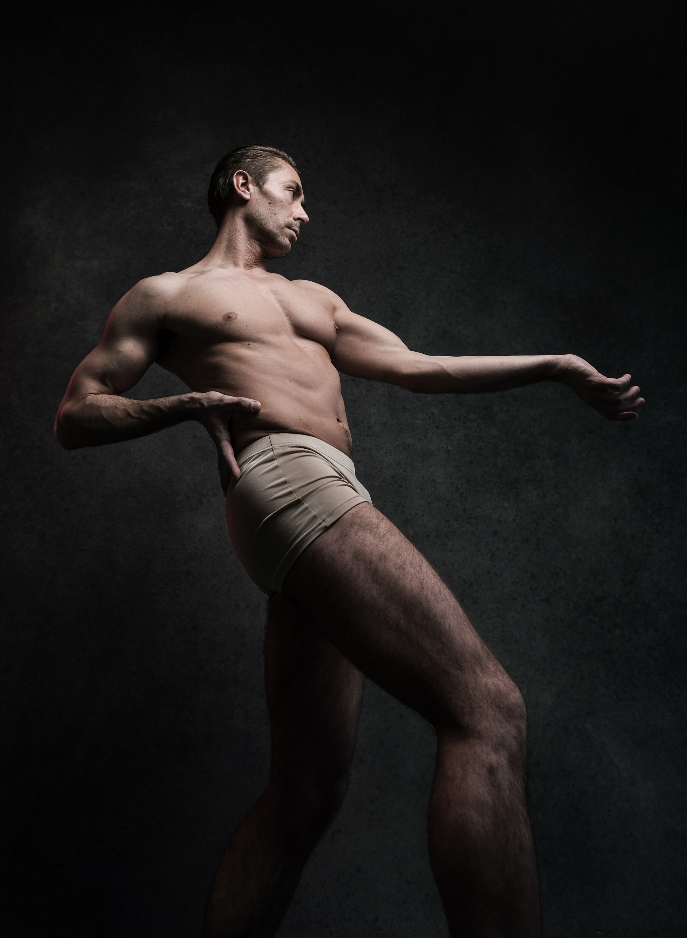 a male dancer is captured midpose against a dark, textured backdrop. his muscular physique is illuminated by studio lighting that highlights his form and the graceful lines of his body. he wears only tan underwear, drawing attention to his physicality and the athleticism of his art. the image conveys a sense of energy and movement, frozen in a moment of artistic expression.