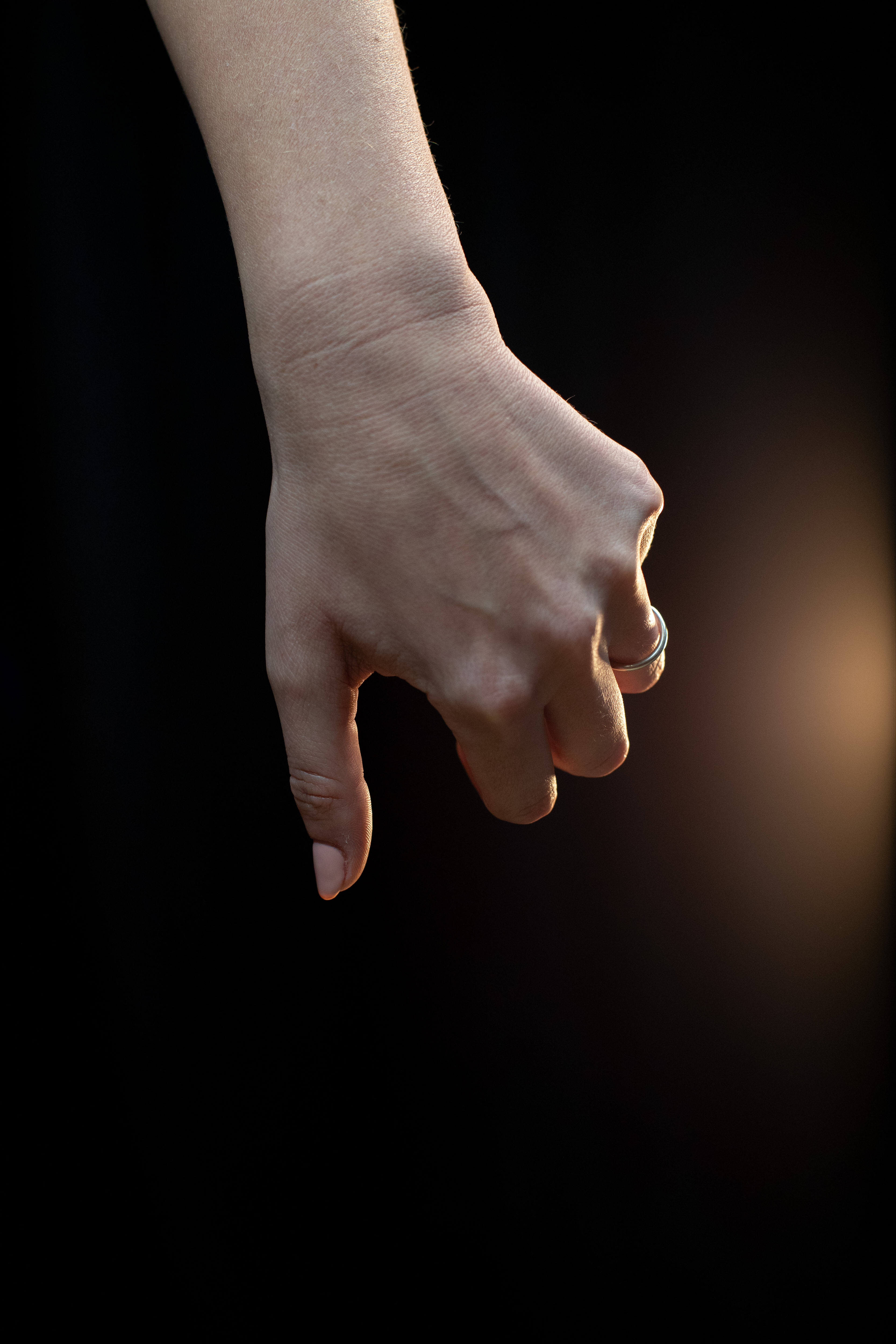 a woman's hand is shown against a black background with a single light source illuminating the hand from the right side. the hand is making a pinching gesture with the thumb and index finger, and the remaining fingers are curled inward. the woman is wearing a simple silver ring on her ring finger.