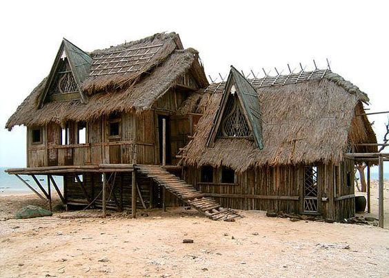 the image shows a dilapidated, rustic dwelling perched on stilts above sandy beach.  the structure appears to be made of wood and has a thick thatched roof. it shows significant signs of age and weathering, with some parts appearing to be falling apart. the house is composed of two connected sections, each with a gabled thatched roof. a weathered wooden ramp provides access to the raised structure. the setting suggests a tropical or coastal environment.  the overall impression is one of abandonment or disrepair, possibly reflecting a state of decay due to exposure to the elements.