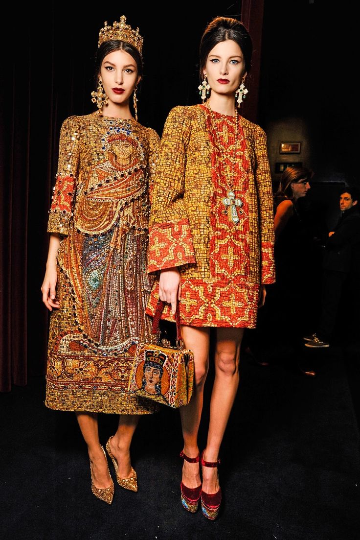 the image shows two models backstage at what appears to be a dolce & gabbana fashion show.  both are wearing elaborate, richly decorated dresses with gold and red mosaiclike patterns.  one model is wearing a longer dress featuring a byzantineinspired religious iconography print; the other is in a shorter, jacketstyle dress. both dresses are richly detailed and luxurious in appearance.  the models are wearing gold jewelry, highheeled pumps (red for one model, gold for the other) and carry ornate handbags. one model also wears a gold crown. the background is dark and suggests the setting is backstage, with other people visible but out of focus. the overall aesthetic is one of opulent, high fashion with religious and historical references.