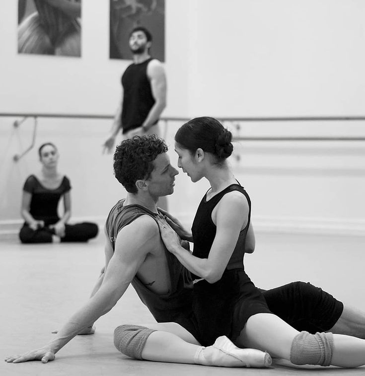 a black and white photo of a male and female dancer, positioned on the floor in a ballet studio. the man is seated with his legs spread wide. the woman is seated with her legs bent with her left leg crossed in front of her right leg. they are looking at each other
