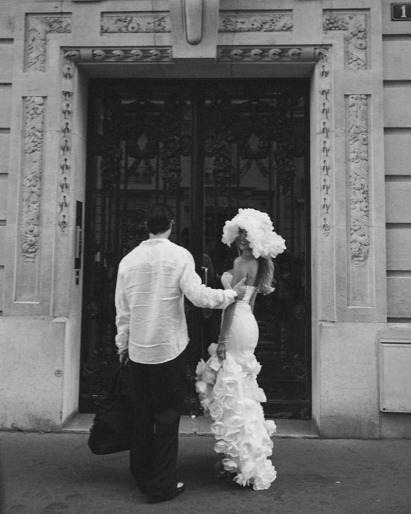a black and white image of a couple in front of a parisian doorway. the woman wears a beautiful white dress and a floral headpiece, while the man is wearing a white buttondown shirt. they look fashionable and elegant, and the architecture of the building adds to the romantic ambiance of the image.