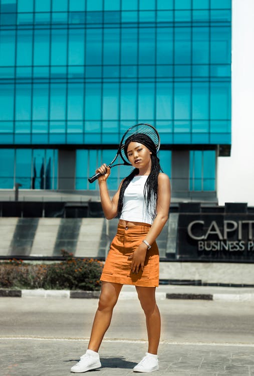 a young black woman with long braids holds a tennis racket over her head. she is wearing a white crop top and an orange mini skirt, along with white sneakers. she is standing outdoors in front of a modern glass building. the overall style is urban and fashionable. the image is bright and sunny, showcasing a summery vibe.