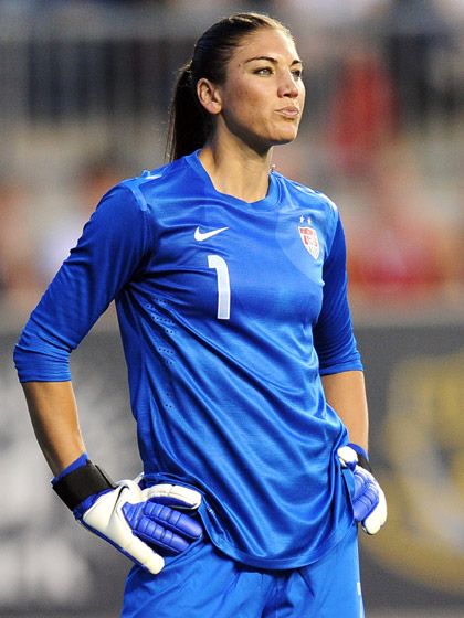 hope solo, goalkeeper for the u.s. women's national soccer team, stands on the field during a game.  she is wearing her blue goalkeeper uniform, with her jersey partially tucked into her shorts. she is wearing her gloves and has her hands on her hips. the background is out of focus, but it is clear that she is on a soccer field with spectators in the stands.
