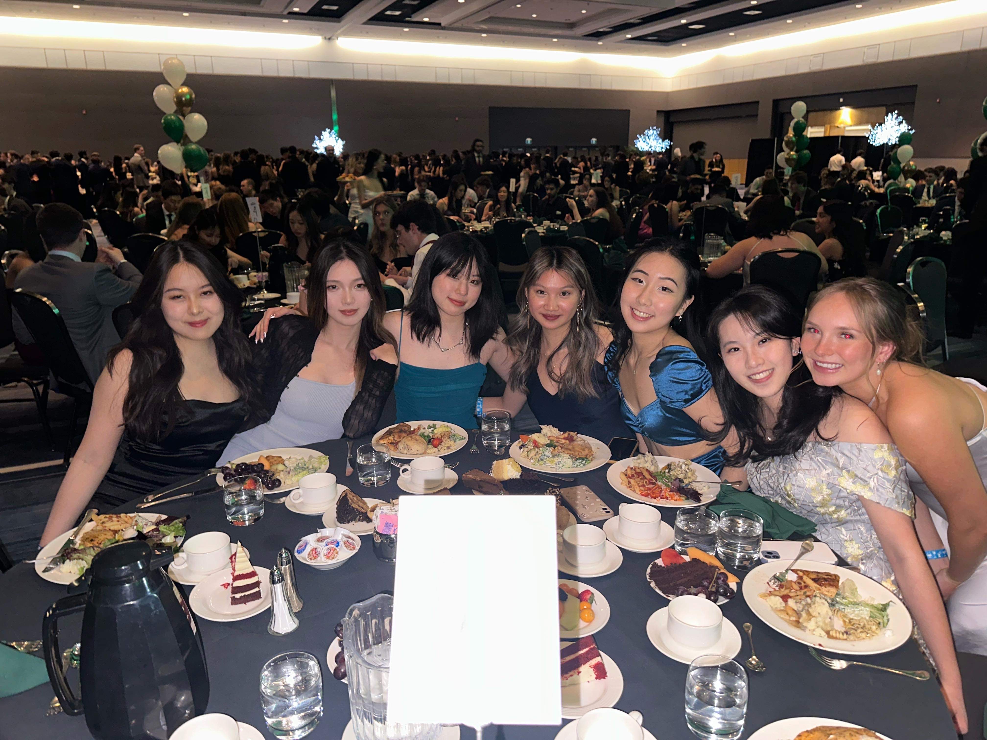 the image shows a group of eight young women sitting at a round table covered with a black tablecloth. they are all dressed in formal attire, likely for a prom or a similar event. the table is laden with food and drinks, including plates of appetizers, main courses, desserts, and beverages.  a white card stands in the center of the table, possibly a table number or a placeholder for a decoration. in the background, a large crowd of people, also dressed in formal attire, can be seen enjoying the event. the ballroom is brightly lit with artificial lighting, creating a festive atmosphere.