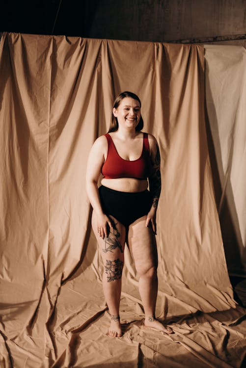 the image shows a confident, plussize woman with tattoos on her legs, smiling gently. she's standing in a studio setting against a light beige backdrop. she's wearing a burgundy bralette and black highwaisted underwear, showcasing a body positive and selfaccepting posture.  the overall mood is one of comfort and selfassurance.