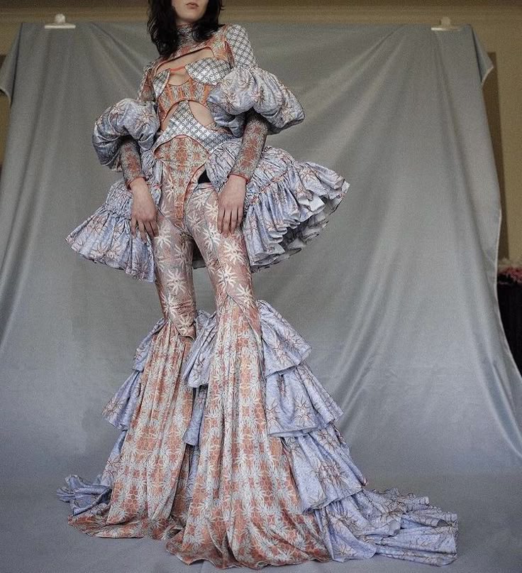 a model poses in an elaborate, avantgarde fashion outfit. the outfit consists of a patterned top with cutout details, voluminous ruffled sleeves, fitted pants with a flared, ruffled bottom that extends into a train. the fabric has a complex, florallike pattern with a blend of peach and bluegrey tones. the model stands in front of a grey backdrop, which helps to emphasize the outfit's unique design.