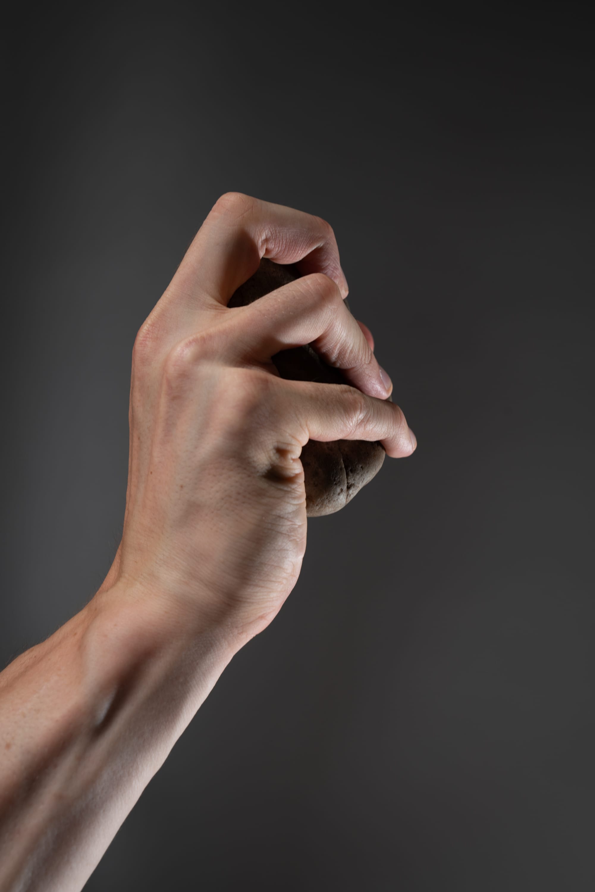 a closeup image of a man's hand tightly gripping a brown potato against a gray background. the potato is positioned vertically, with the man's fingers curled around its circumference. the image showcases the hand's strength and the texture of the potato's skin. the lighting is subtle, highlighting the contours of the hand and the potato.