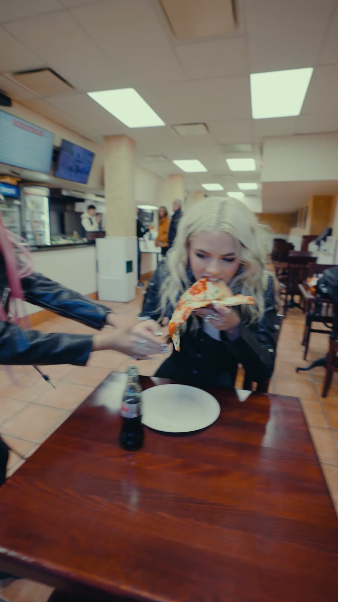 a woman with blonde hair is eating pizza inside a restaurant. there's a table with a plate and a bottle of soda. another person is visible on the left side of the frame, with their hand partially reaching towards the woman. the restaurant has an indoor setting with visible food service areas in the background.