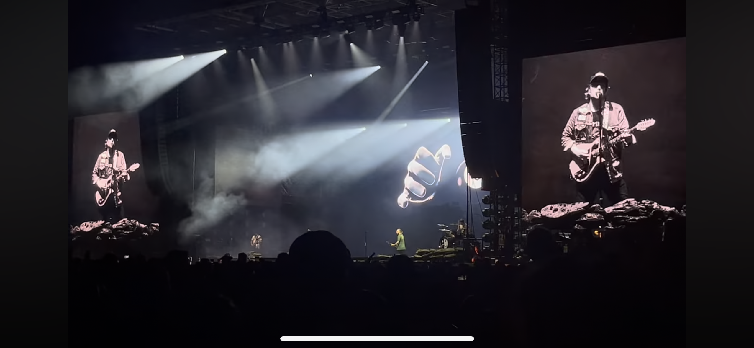 a concert scene captured from the audience, showcasing the stage bathed in spotlights, with a rock band performing. the stage is dimly lit, with the performers standing in the bright light. the crowd is partially visible in the foreground, a blur of heads and silhouettes. the band is playing to a large crowd with the energy of a live concert experience.