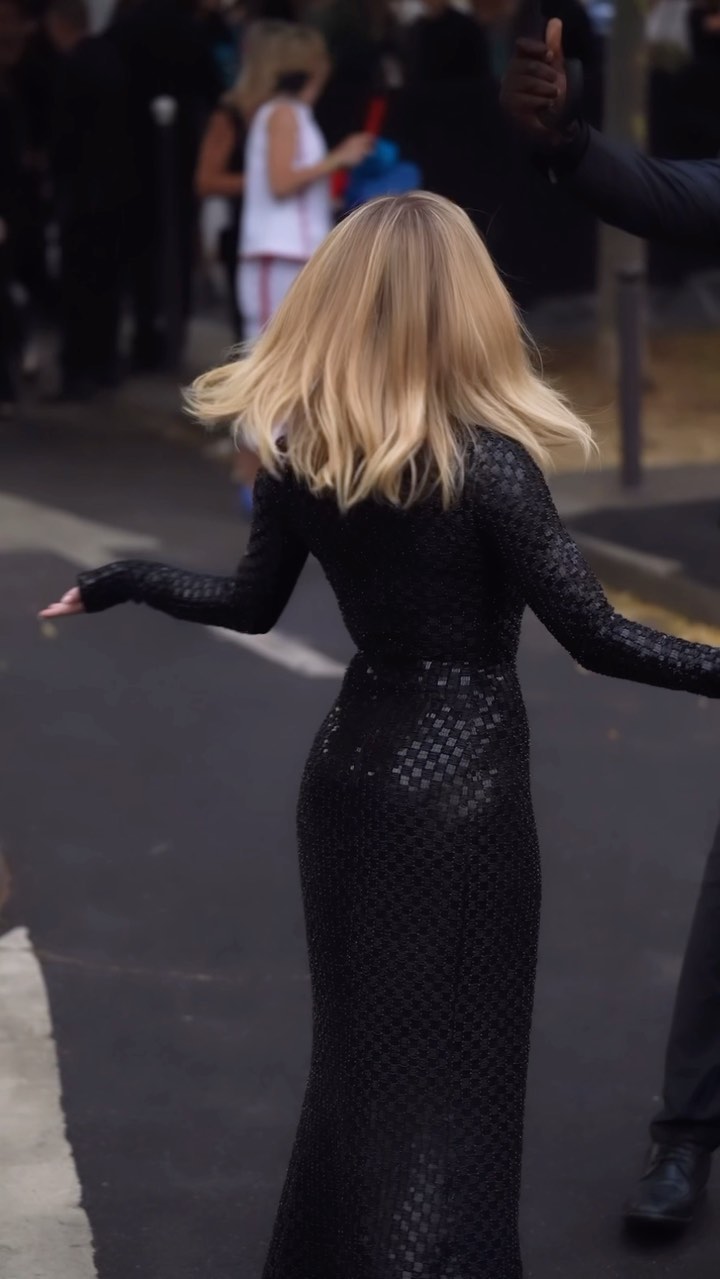 a blonde woman in a long, black, textured dress walks down a city street. the dress has long sleeves and a figurehugging silhouette.  she is moving slightly, suggesting motion.  the background is blurred but shows other people and some urban details, suggesting the photo was candidly taken. the woman's hair is loose and flows behind her.  a portion of a person's arm and dark clothing is visible in the upper right corner, suggesting someone is assisting or walking with her. the overall mood is stylish and elegant.