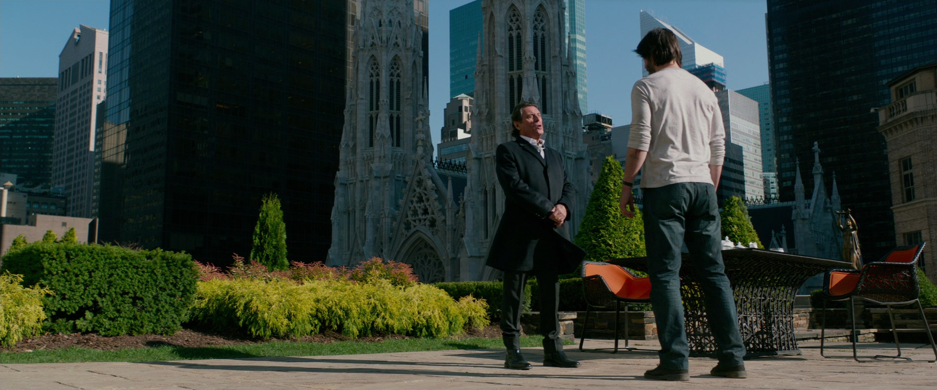 two men are standing on a sidewalk in front of a skyscraper in the city, they are looking at each other and talking. one man is wearing a suit and the other man is wearing a white tshirt and jeans.  there are some buildings in the background.