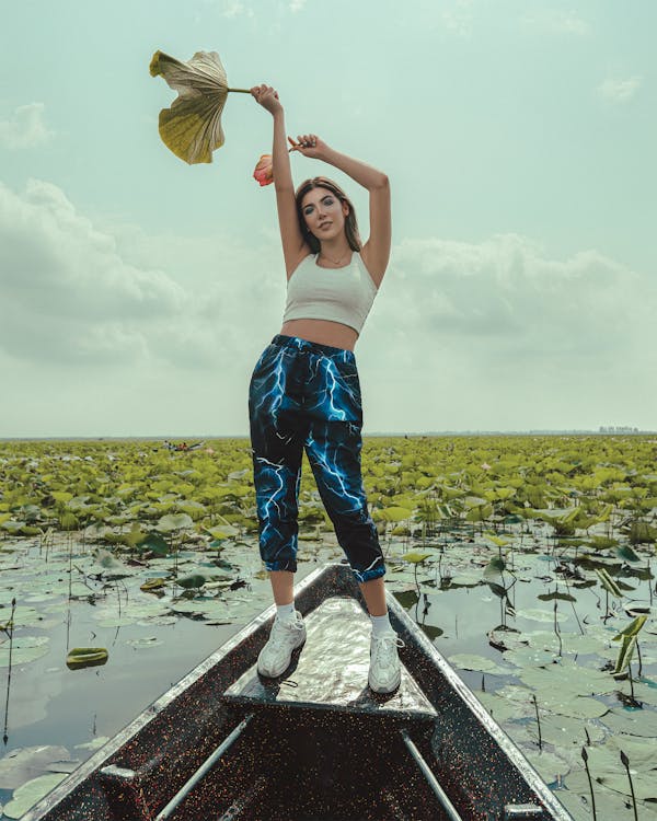 a young woman with long brown hair is standing in a small boat on a calm lake filled with vibrant green lotus plants. she is smiling and holding a large, dried lotus leaf above her head.  she's wearing a white crop top and stylish blue pants with a lightning bolt print, along with white sneakers. the scene is tranquil and sunny, evoking a feeling of peace and summer vacation. the sky is partly cloudy.