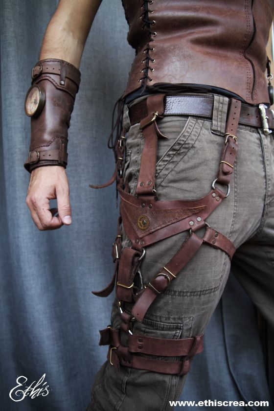 the image showcases the lower torso and leg of a person modeling intricate steampunkinspired leather attire.  the focus is on a complex harness system crafted from brown leather with metal buckles and rivets. the harness wraps around the model's thigh and connects to a belt, adding a rugged and functional aesthetic. the model also wears cargo pants and a leather wrist cuff with a clock embedded in it. the backdrop is a muted blue, drawing attention to the craftsmanship of the leatherwork.