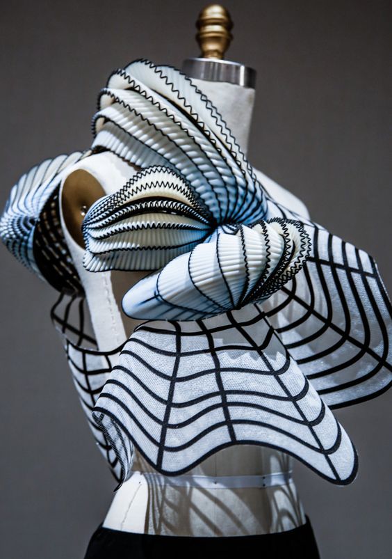 a closeup shot of a white and black geometric dress, showcasing a unique 3d design with intricate pleats and patterns. the dress appears to be made of a lightweight, translucent fabric and features a futuristic, avantgarde aesthetic. it is displayed on a mannequin against a plain gray background. the lighting highlights the dress's intricate details, creating a captivating and artistic composition.
