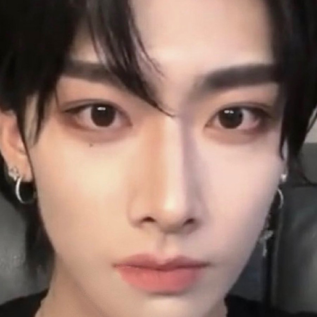 a closeup selfie of a young asian man with styled black hair and makeup. he is looking directly at the camera with a neutral expression. the lighting is soft, highlighting his smooth skin and the details of his makeup. he is wearing small silver hoop earrings. the overall aesthetic leans towards a trendy, androgynous kpop idol look.