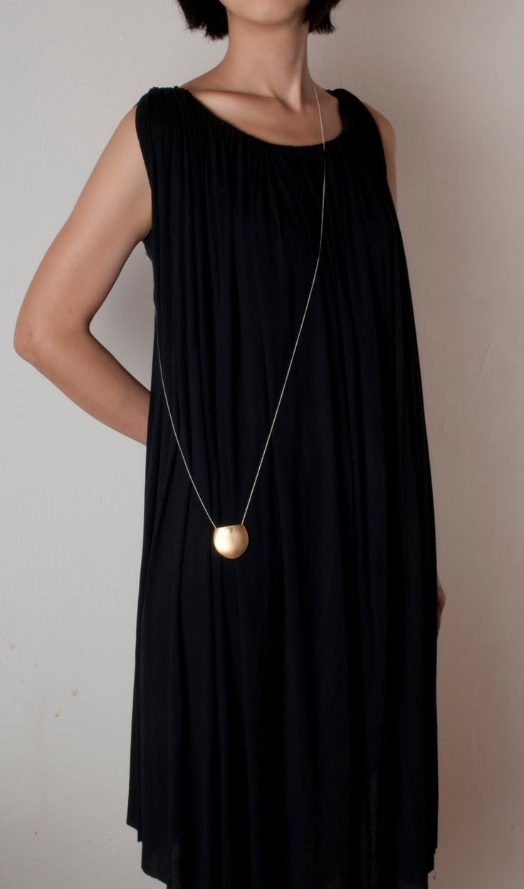 a woman is wearing a black dress and a gold necklace with a round pendant. the dress is sleeveless and has a loose, flowing fit. the necklace is long and thin, and the pendant is a simple round shape. the woman is standing against a plain background.