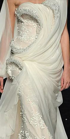 a bride wearing a white gown adorned with intricate lace and beading. the dress has a flowing, dramatic silhouette and features a high neckline and long sleeves. the bride is posing with her head tilted slightly, showing off the dress's detailing. the image is captured in a studio setting and has a soft, romantic feel.