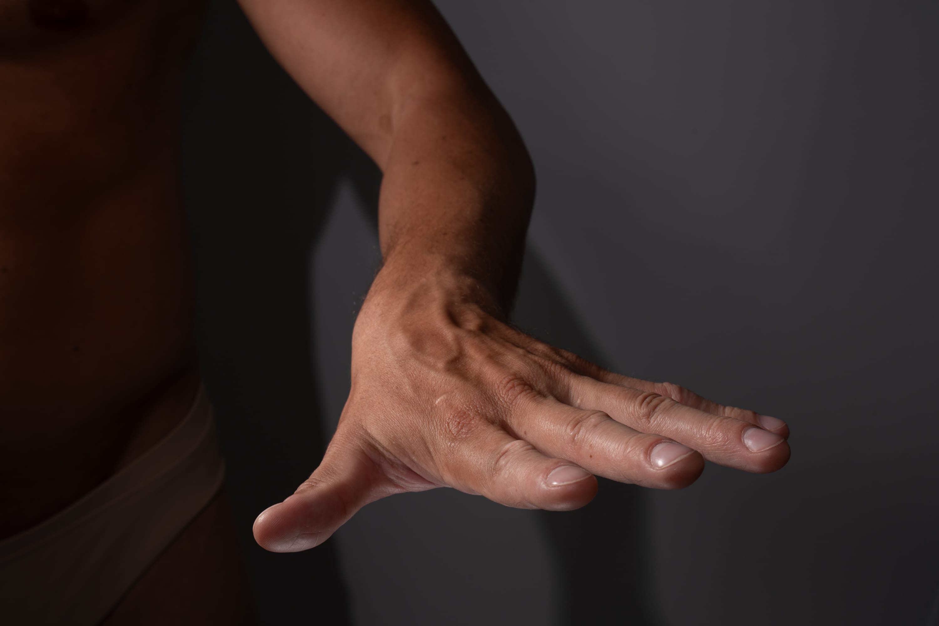 a closeup image of a muscular man's outstretched hand and arm against a dark gray background. the image emphasizes the details of the man's hand, showcasing the veins and muscle definition. the studio lighting creates a dramatic contrast, highlighting the contours of his body.