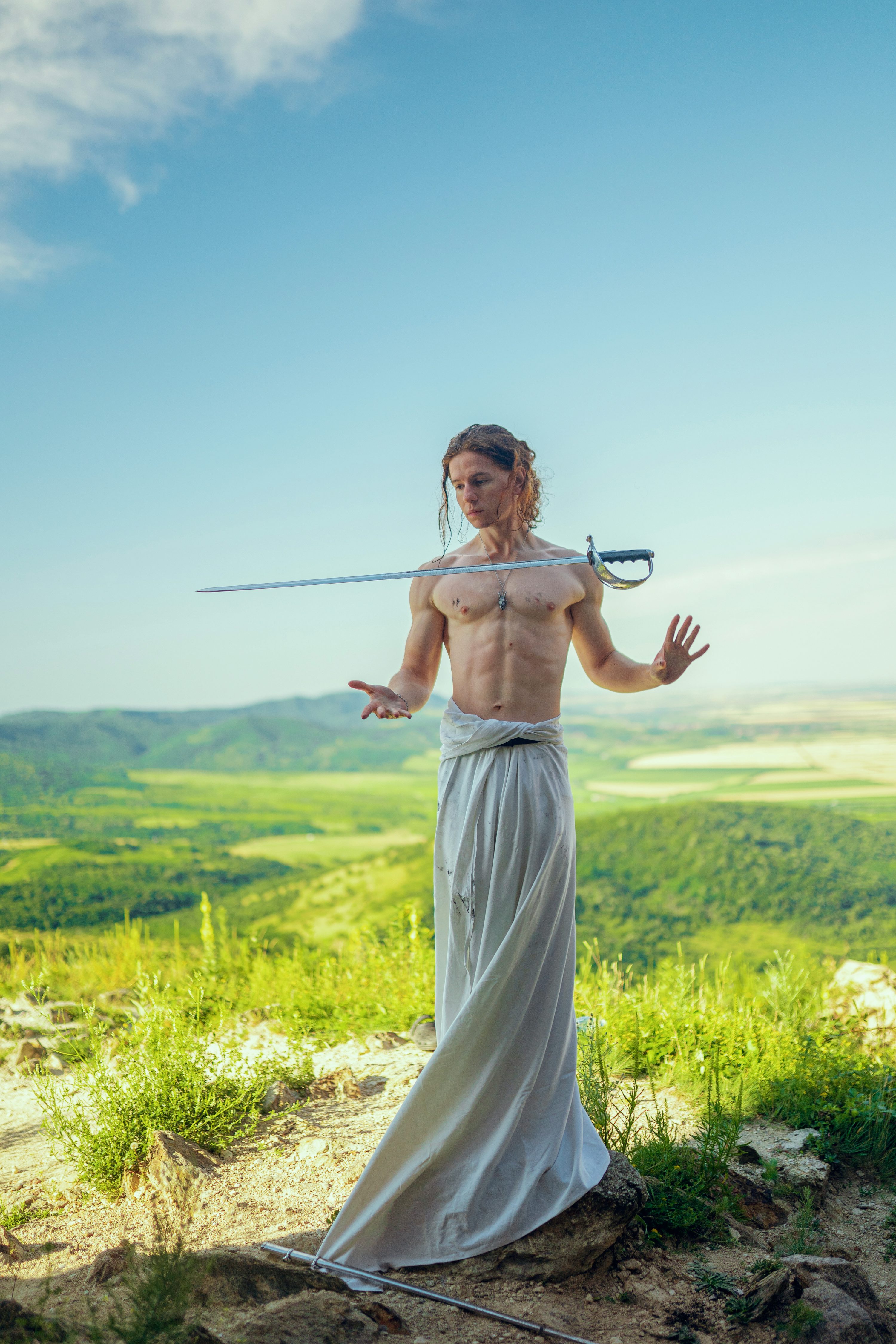 a shirtless man with long hair stands on a hilltop, balancing a sword across his shoulders. he wears a long, flowing white robe. the landscape behind him is a beautiful, green valley under a bright, partly cloudy sky. the image is artistic and evocative, possibly suggesting themes of strength, balance, or spiritual connection with nature.