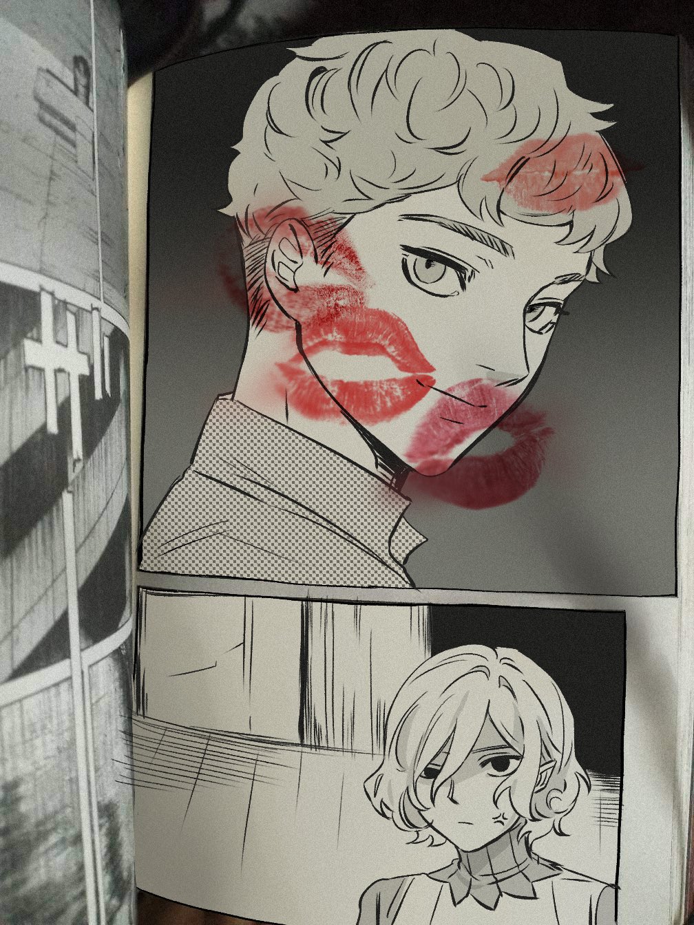 the image shows an open manga or comic book.  the main focus is on two panels. the top panel features a young man with short, lightcolored hair, his face covered in numerous red lipstick kiss marks.  the style is predominantly black and white, with the red kiss marks providing a striking contrast. the bottom panel displays a young woman with short, light hair. she has a more subdued expression compared to the energetic implication of the man's panel. the overall impression is one of romance or a romantic moment, suggested by the kiss marks on the man's face. the left edge of the photograph shows the open pages of what appears to be another section of the comic book.